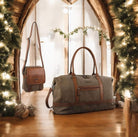 Two bags, a gray duffel bag and a brown crossbody bag, in front of a decorated Christmas tree.